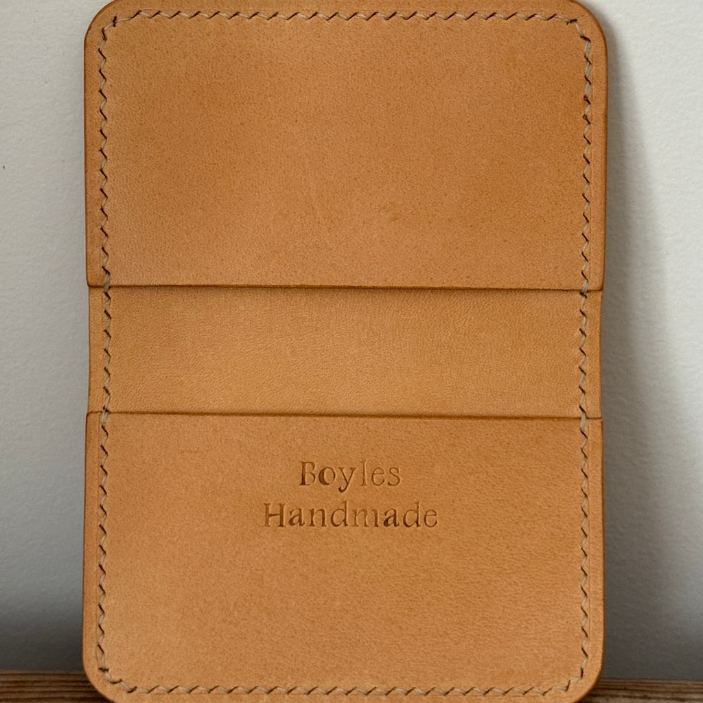 A tan leather card holder wallet with visible stitching and the debossed text 'Boyles Handmade' on the front, standing upright on a wooden surface against a light gray wall.