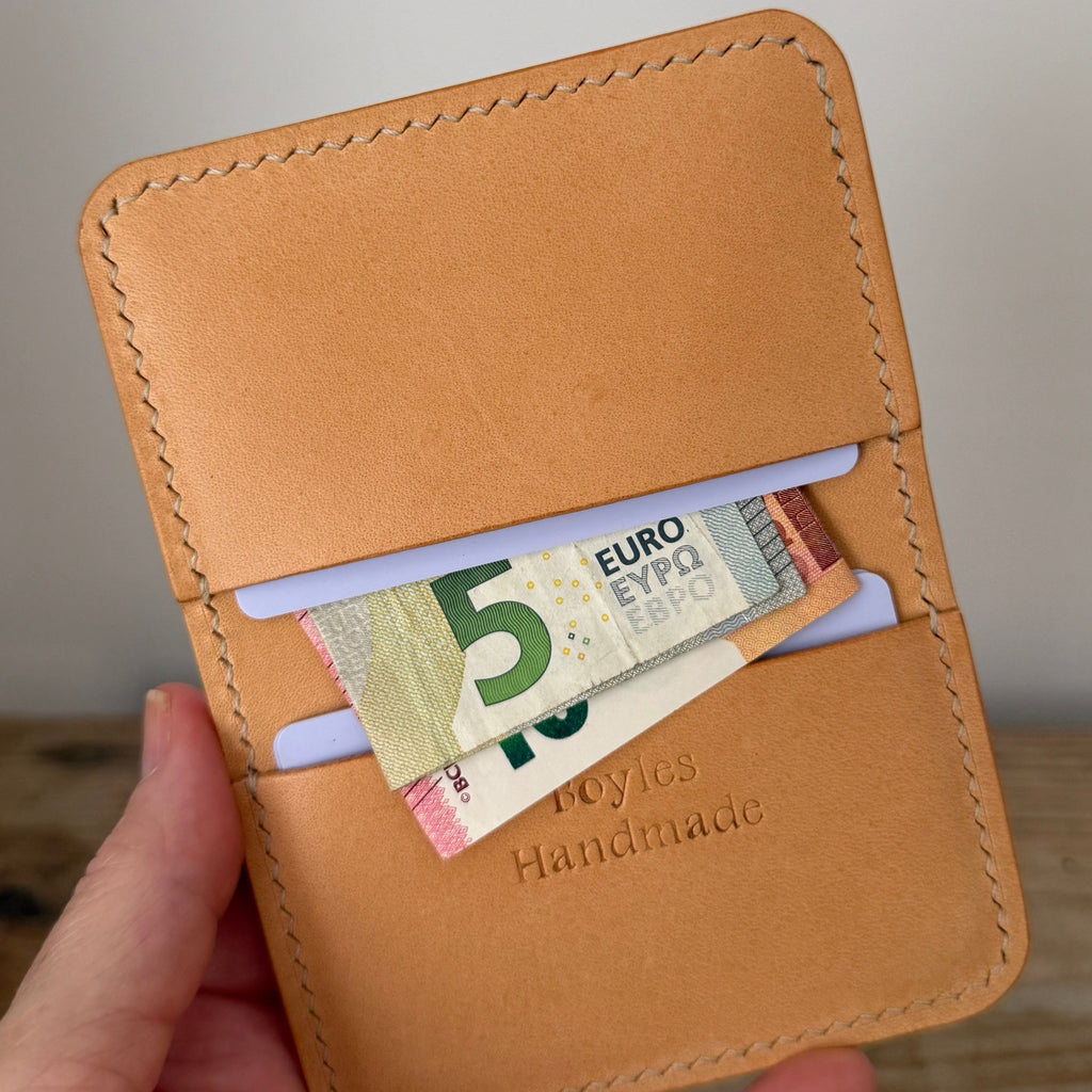 A hand holding a tan leather card holder wallet with a 5 Euro banknote tucked into one of its slots. The wallet features visible stitching, a debossed 'Boyles Handmade' logo, and is photographed against a neutral light gray wall and wooden surface background.