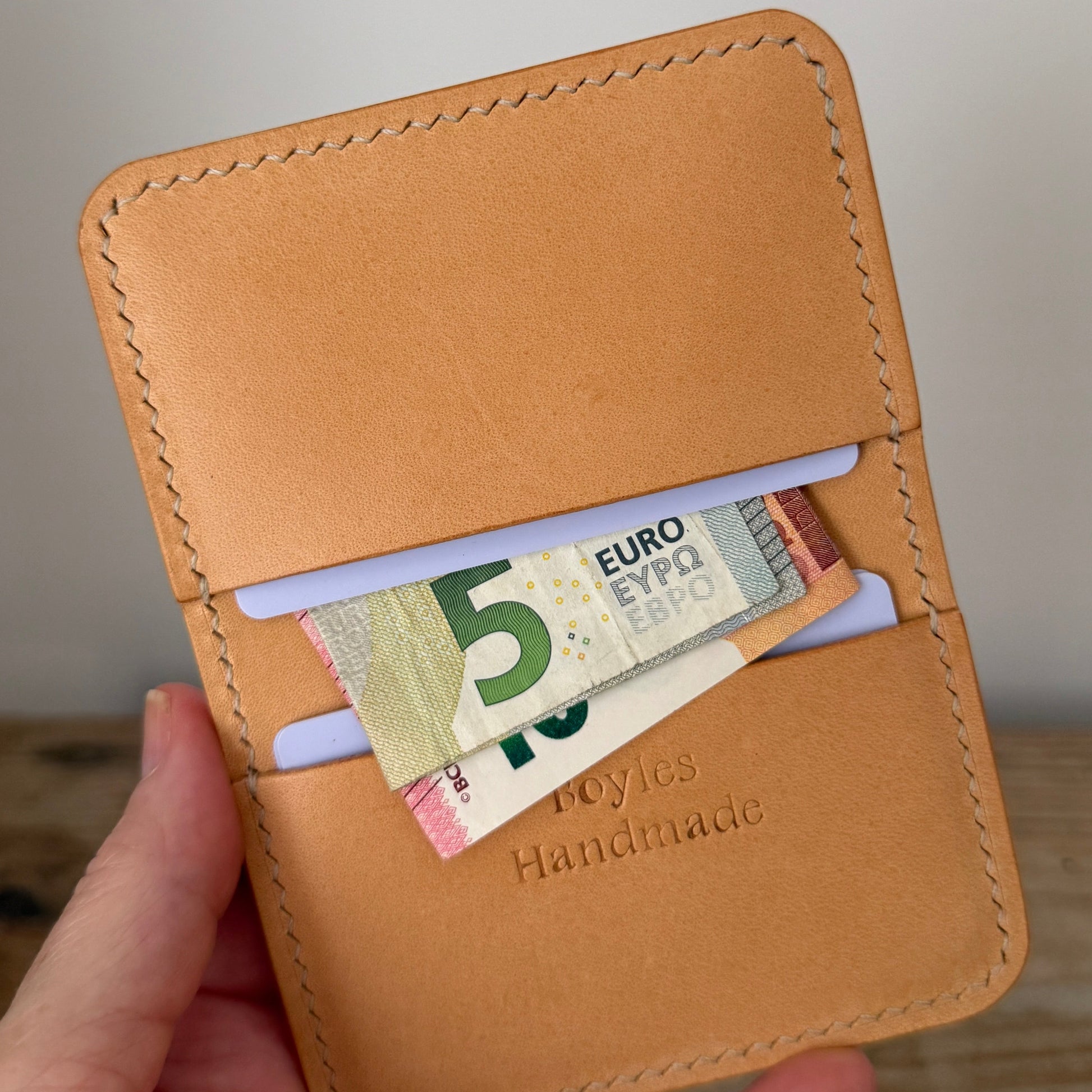 A hand holding a tan leather card holder wallet with a 5 Euro banknote tucked into one of its slots. The wallet features visible stitching, a debossed 'Boyles Handmade' logo, and is photographed against a neutral light gray wall and wooden surface background.
