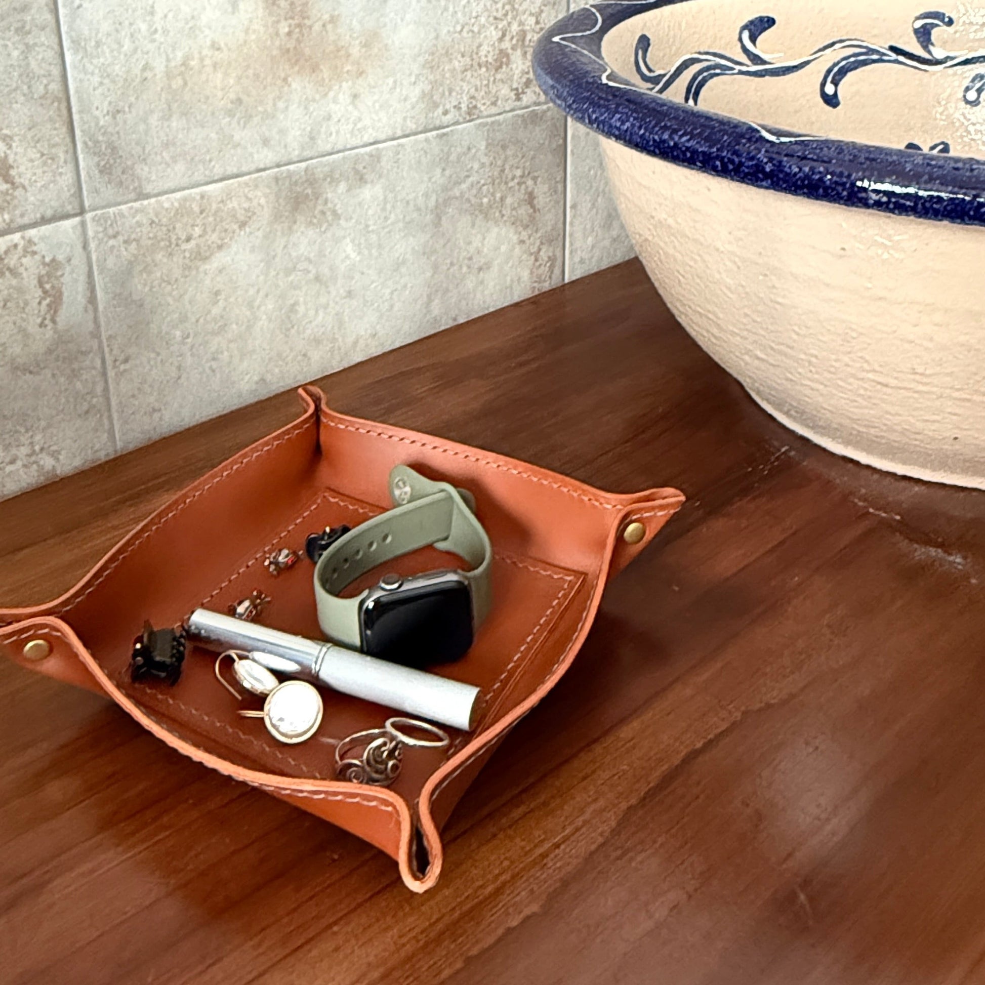 A brown leather jewelry tray holding a smartwatch, keys, and small accessories, placed on a wooden countertop next to a ceramic sink with a blue floral rim and a stone lion faucet against tiled walls.