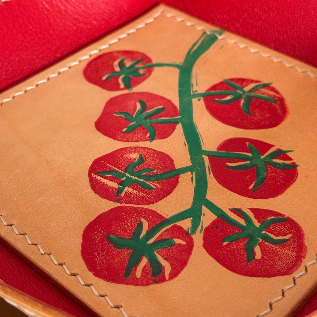 A close-up of a tan leather tray or box interior featuring a printed red tomato vine design with green stems, surrounded by a bright red leather rim with visible stitching.