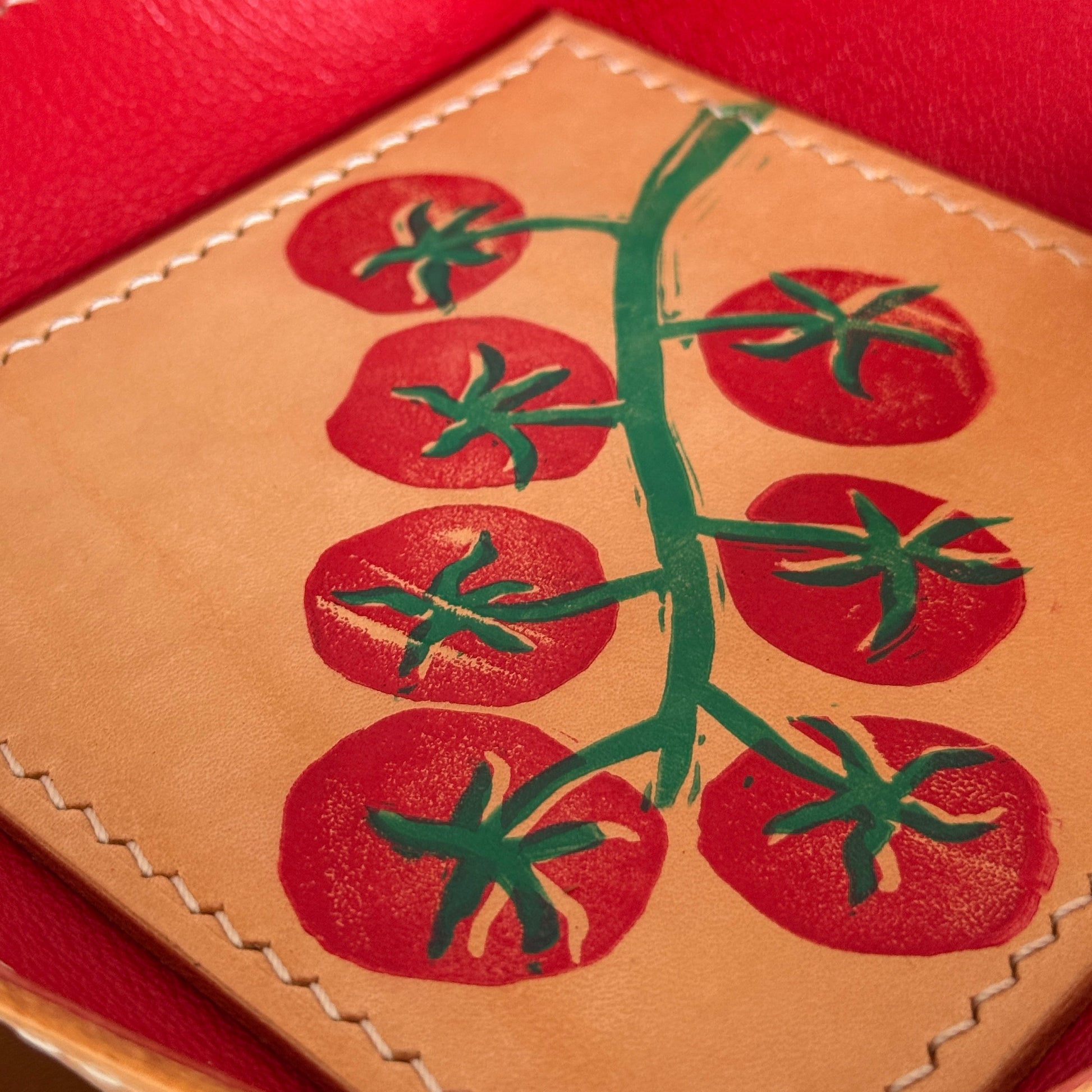 A close-up of a tan leather tray or box interior featuring a printed red tomato vine design with green stems, surrounded by a bright red leather rim with visible stitching.